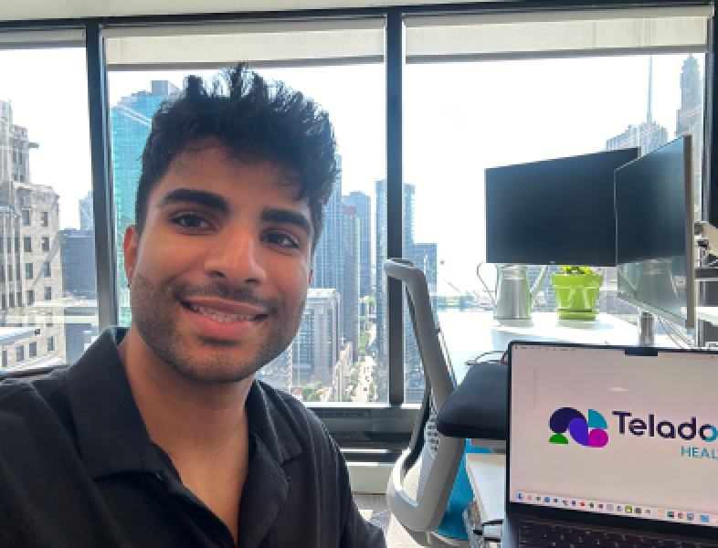 An intern smiling into the camera, sitting in an office with a city skyline in the background and his computer open.