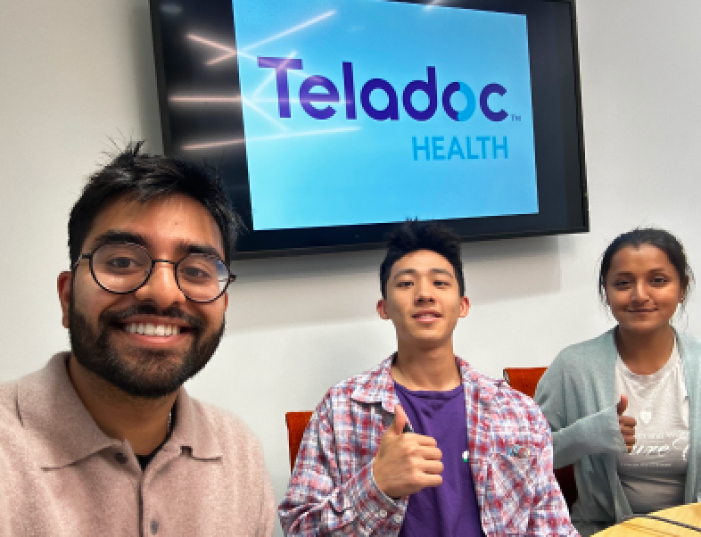 A group of interns in the office with a TV in the background showing the Teladoc Health logo.