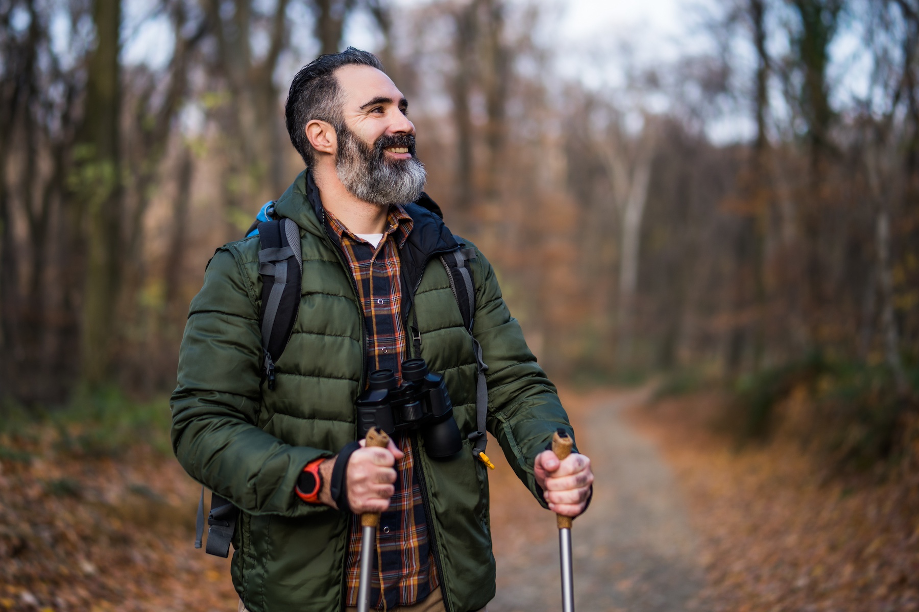 Man hiking through a forest trail with trekking poles and binoculars, enjoying nature.