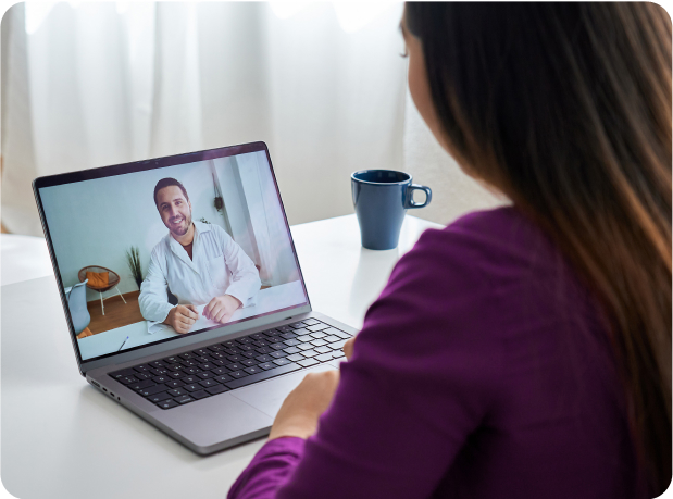 Over-the-shoulder shot of a woman on a video call with a doctor, wearing a purple shirt.