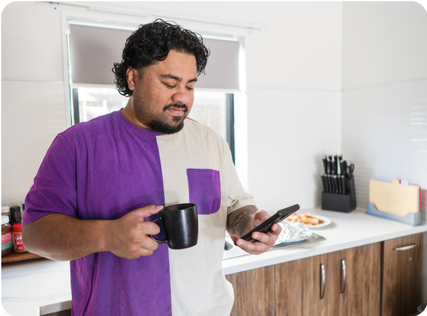 Man smiling at his phone while holding a cup of coffee in his kitchen, wearing a purple and white shirt.