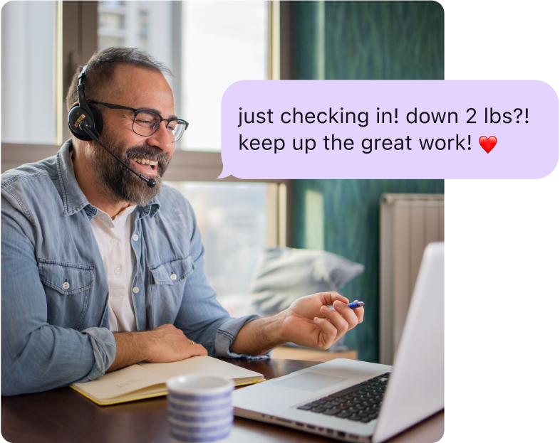 Man with a headset smiling and talking to his computer, with an overlay of a coaching message reading 'Just checking in! Down 2 lbs?! Keep up the great work!'"